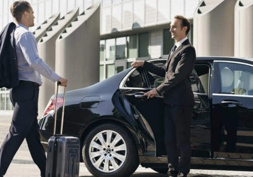 A well-dressed chauffeur in a black suit holds open the rear door of a luxury black sedan for a businessman carrying a rolling suitcase and a jacket over his shoulder, outside a modern building.